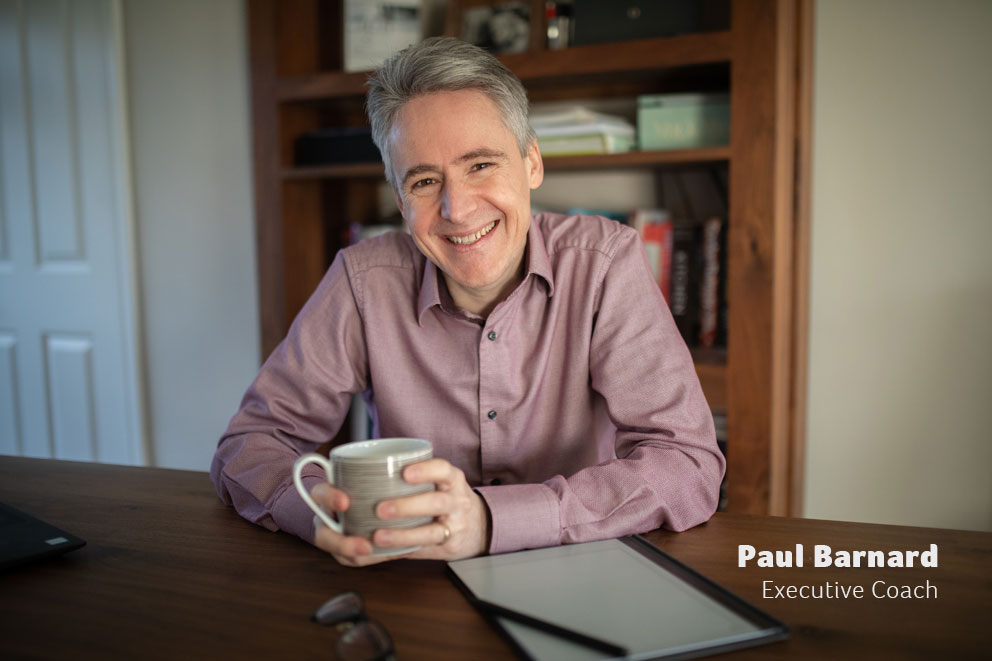 Executive Coach, Paul Barnard sitting at a desk smiling with a hot drink.